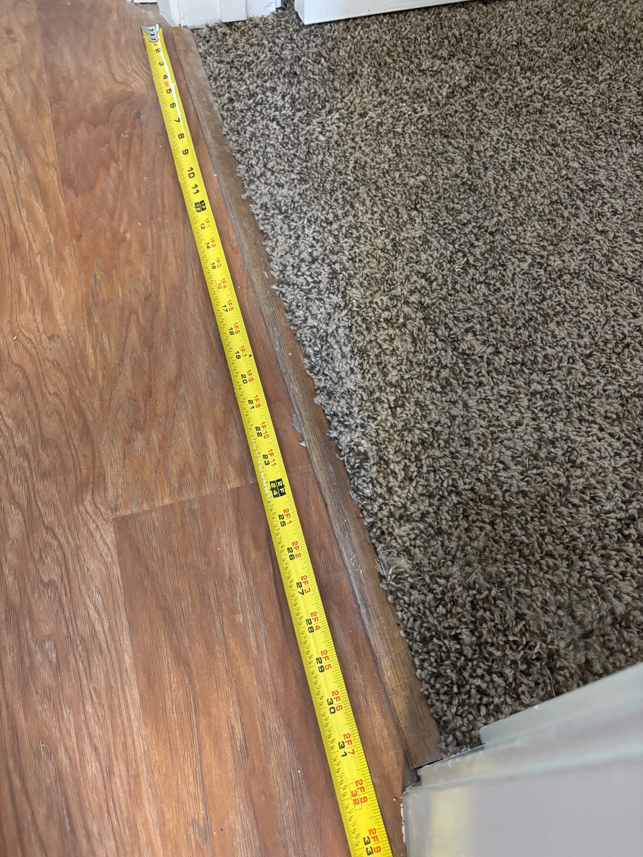 A measuring tape laid out on a hardwood floor next to a carpet, indicating the boundary between the two surfaces.