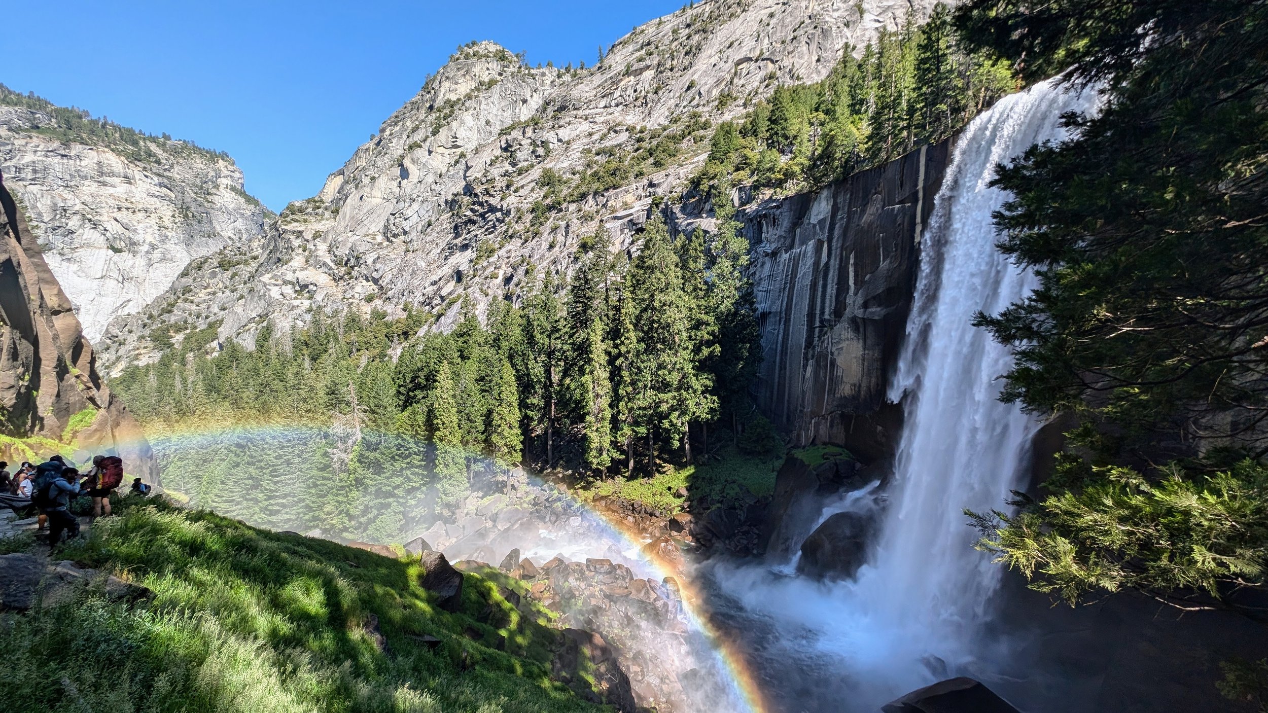 Hiking group near a waterfall with a rainbow in a forested mountain landscape.