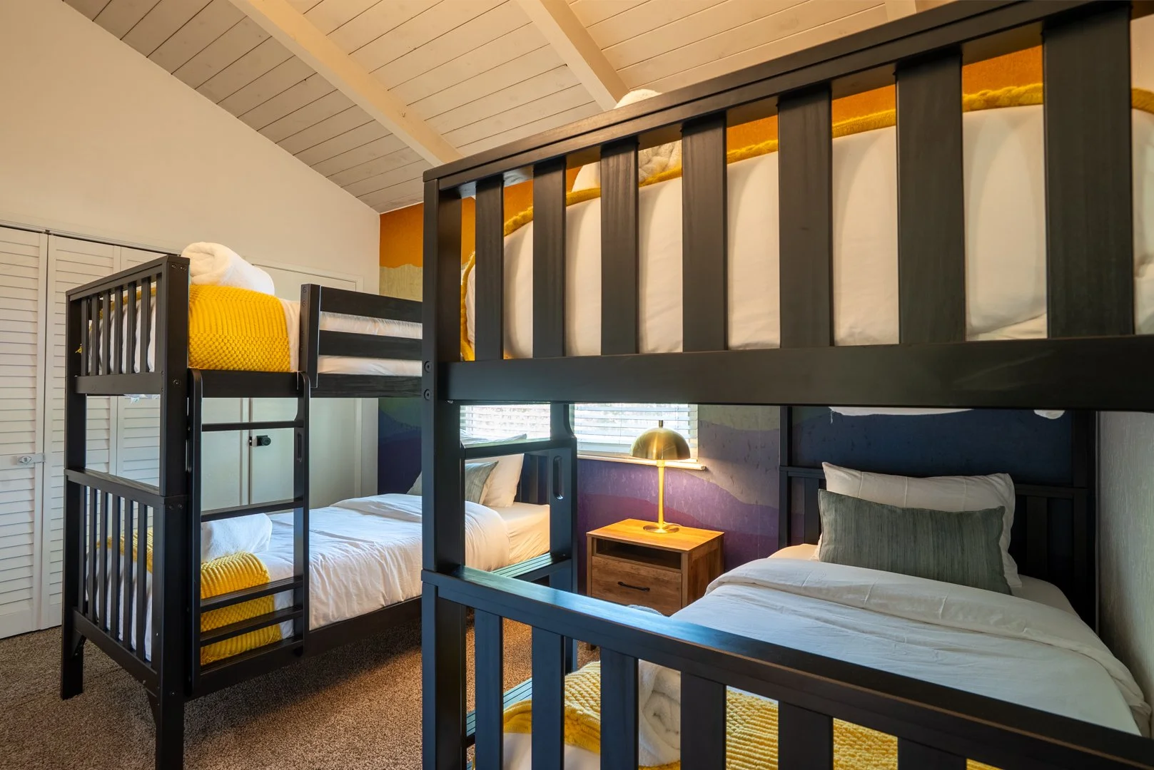 Room with two bunk beds, a nightstand with a gold lamp between them, and a window with blinds. The beds are made with white linens and pillows, and the top bunk has yellow blankets. The room has a wood-paneled ceiling and a multicolored wall behind t