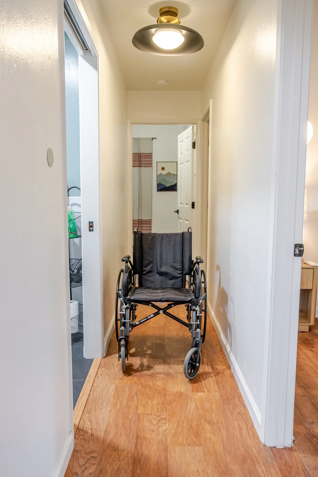 A hallway with a wheelchair in the center, wooden floors, cream-colored walls, and a ceiling light fixture.