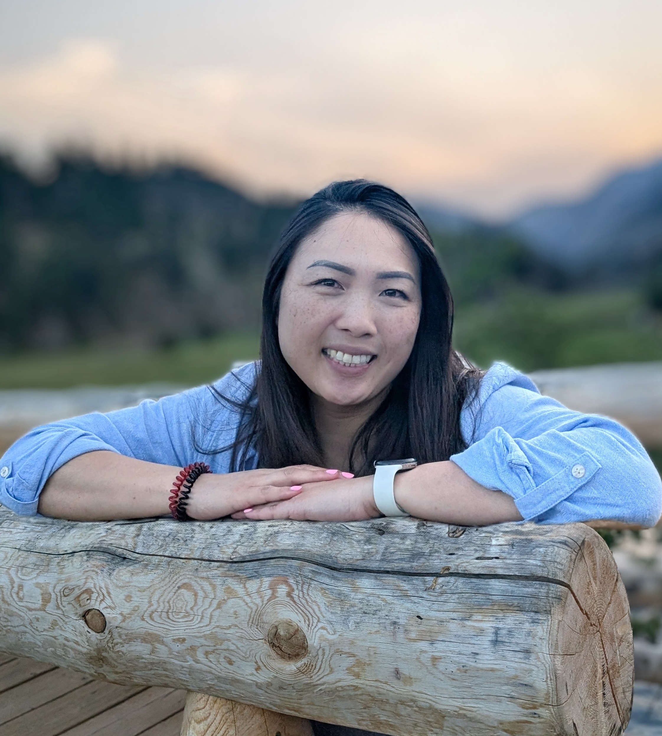 A woman with dark hair smiling, resting her arms on a large, weathered wooden log outdoors, with a blurred natural background and a soft sunset sky.