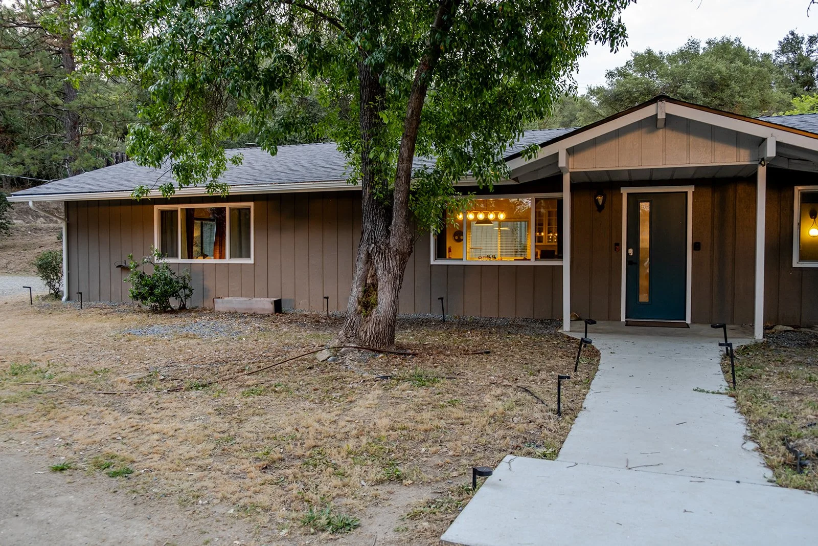 A single-story house with brown vertical siding, a dark blue front door, and a porch light. A concrete walkway leads to the entrance, with small landscape lights along the path. There are trees and dry grass in the front yard, and the house interior 