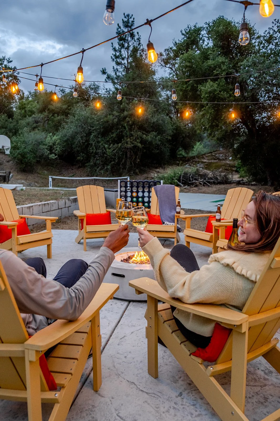 Two people sitting around a firepit, drinking wine and toasting with glasses, outdoors in the evening with string lights overhead and wooded background.