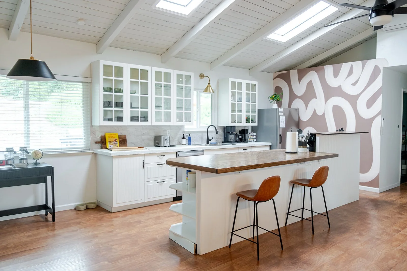 Modern kitchen with white cabinets, a wooden island with two bar stools, and decorative wall art with abstract patterns, illuminated by skylights.
