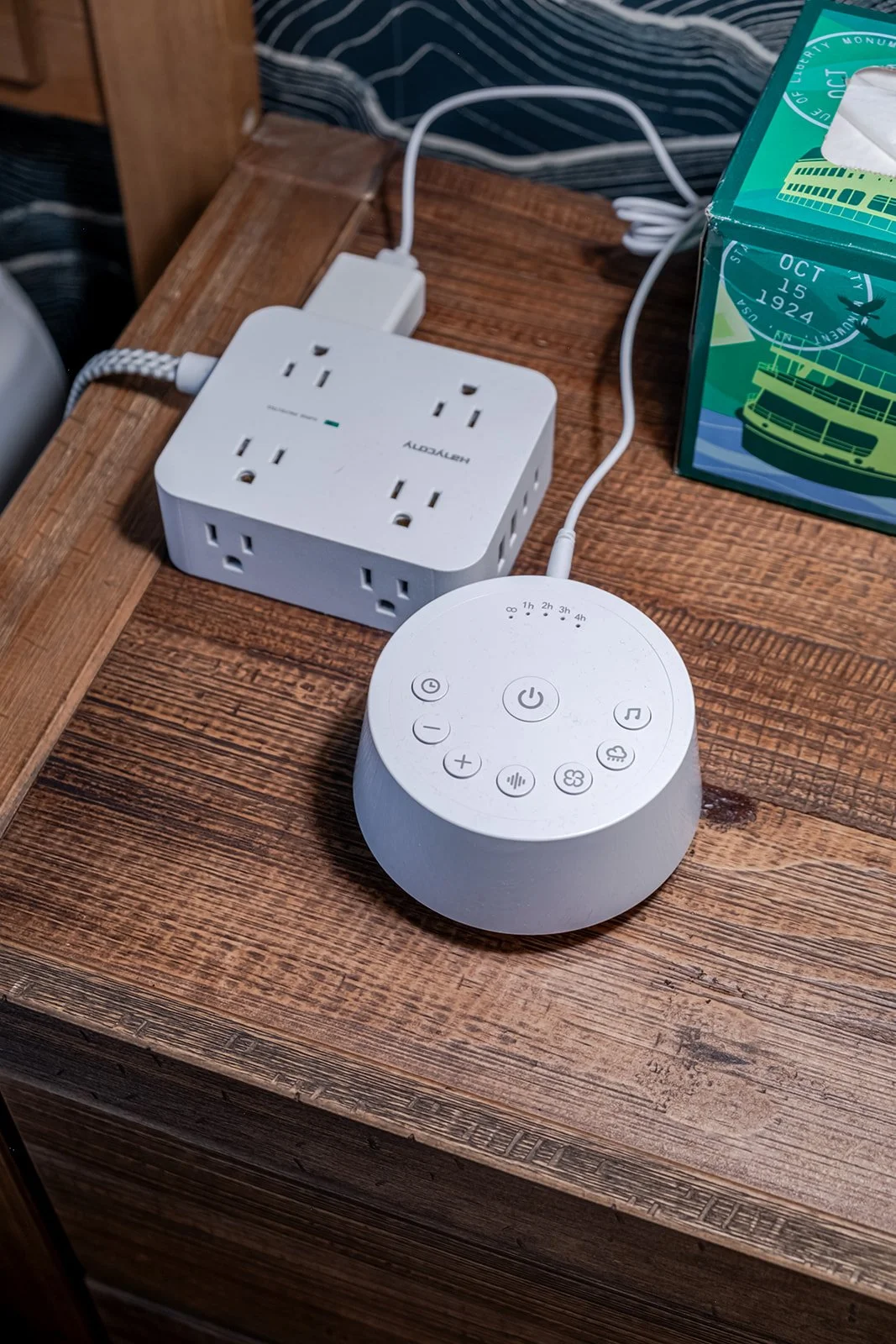 A smart speaker with a circular design, digital controls on top, plugged into an outlet on a wooden table.