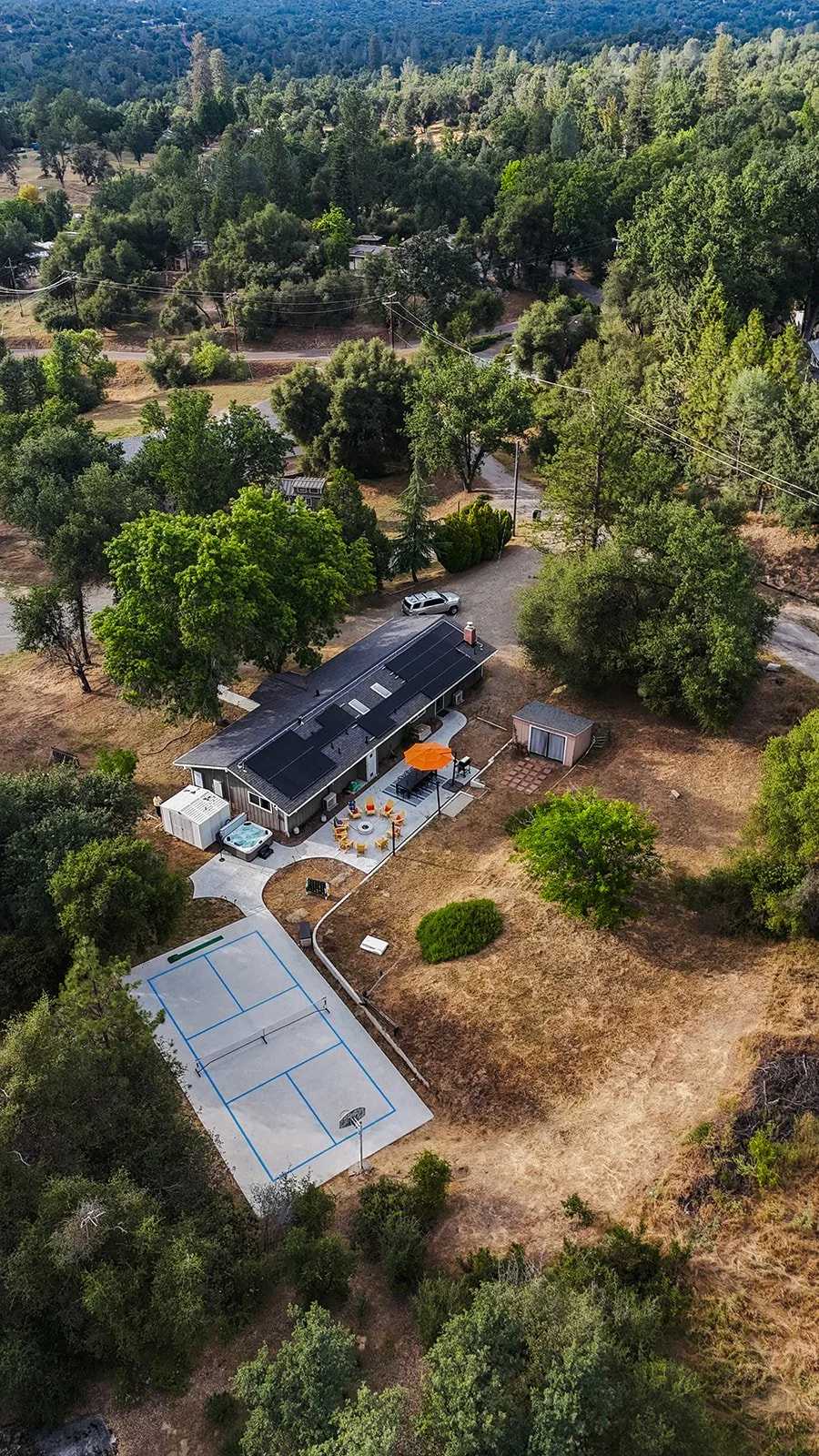 Aerial view of a backyard with a house, outdoor seating area with orange umbrella, a small shed, a basketball court, and lush green trees in a suburban neighborhood surrounded by hills.