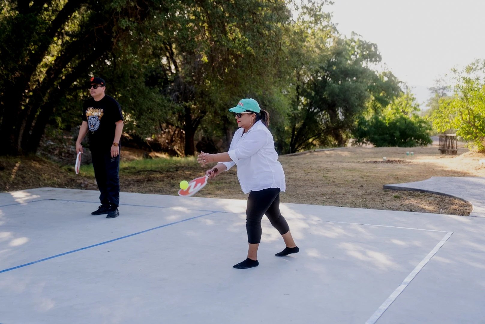 Two people playing pickleball on an outdoor court surrounded by trees. The woman in the foreground is wearing a white top, black pants, a blue cap, and sunglasses, preparing to hit a yellow pickleball with a paddle. The man in the background is wearing a black shirt, black pants, and a black cap, holding a paddle.