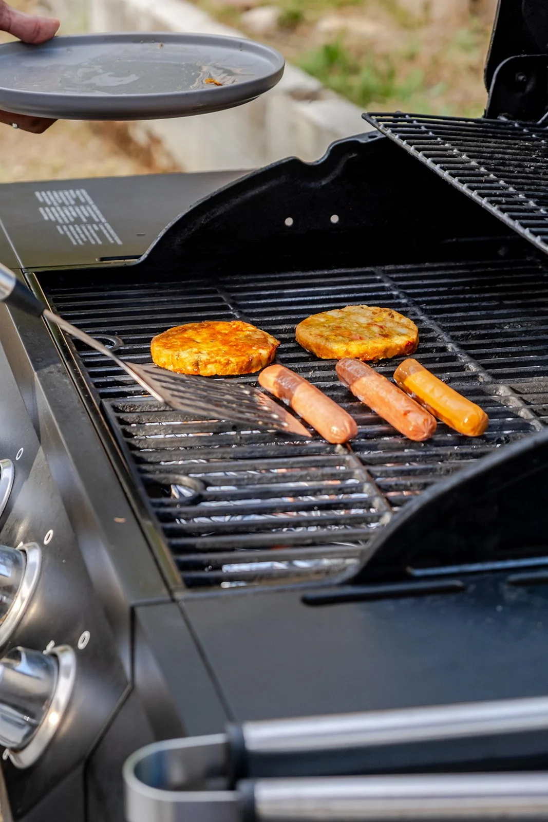 On a barbecue grill, two veggie patties and three hot dogs are cooking. A person is holding a plate near the grill, ready to serve.