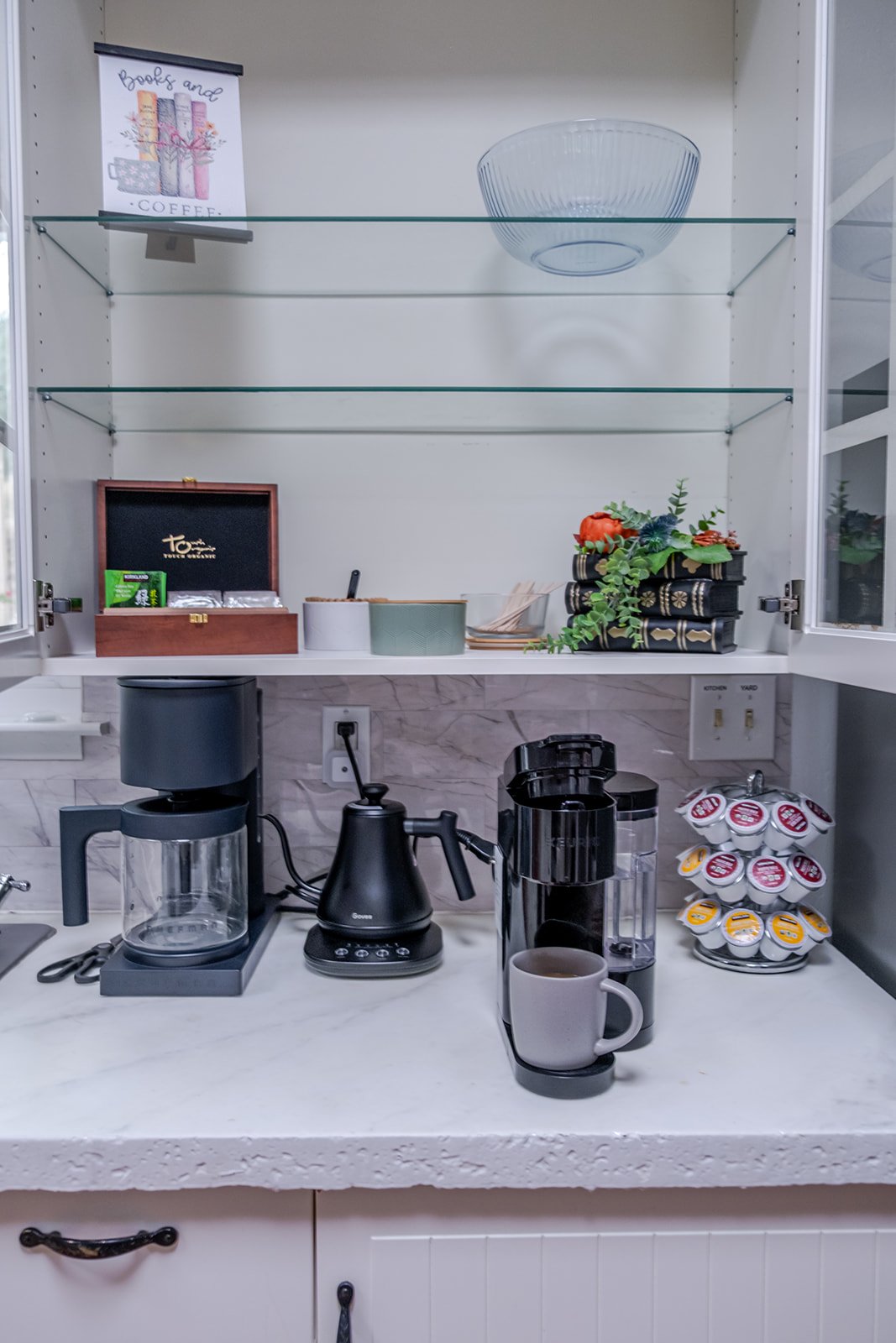 Kitchen countertop with coffee maker, coffee pods, coffee kettle, glass coffee pot, mug, and open cabinet with glass bowl, books, and decorative plants.