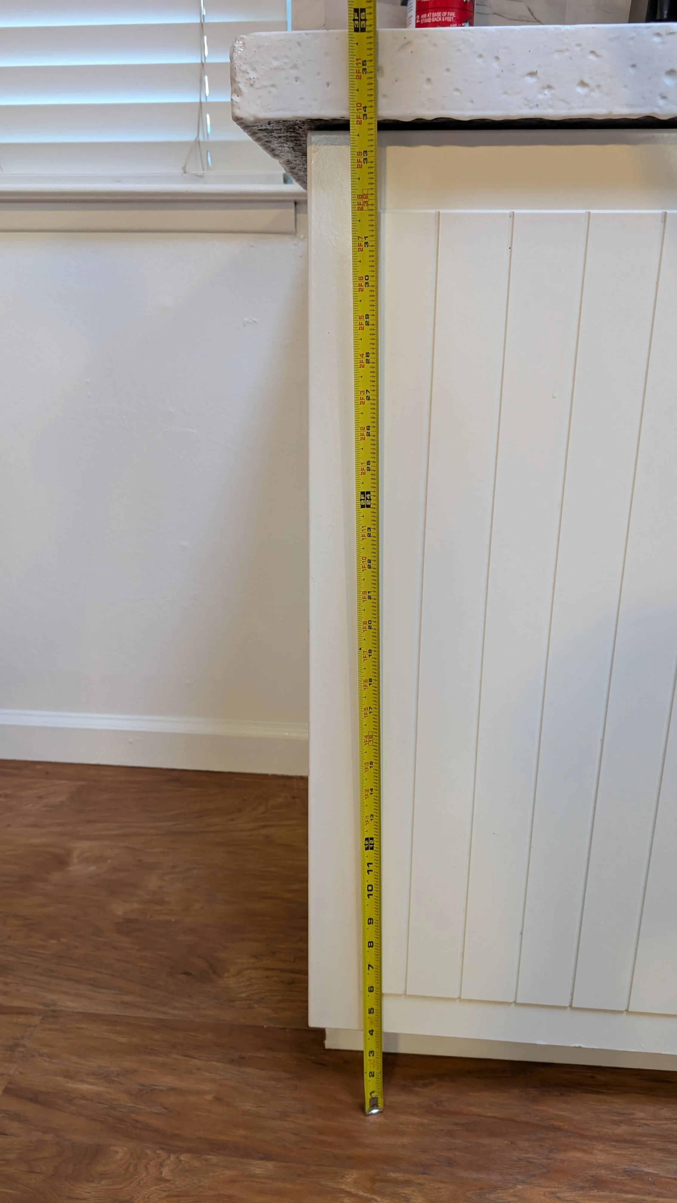 A close-up of a tape measure standing vertically next to a kitchen or bathroom cabinet, showing the height measurement in inches.