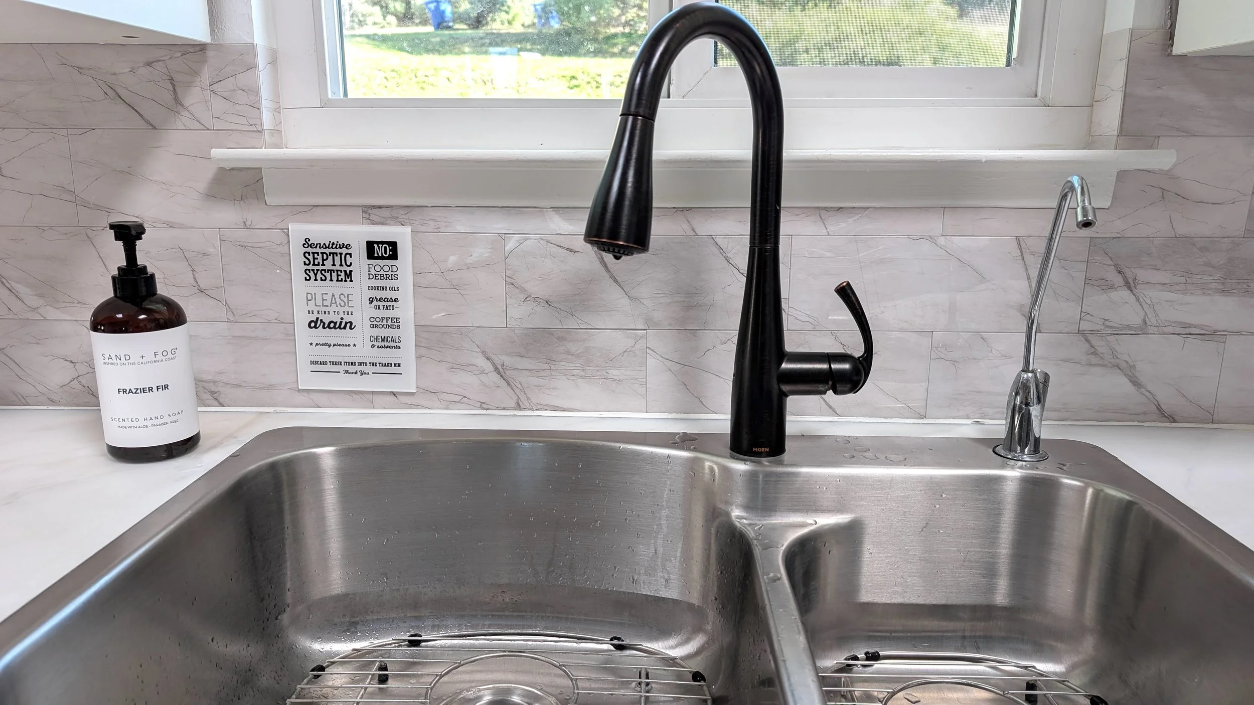 A touchless kitchen sink with black faucet, soap bottle, hand-written note about septic system, and window above the sink.