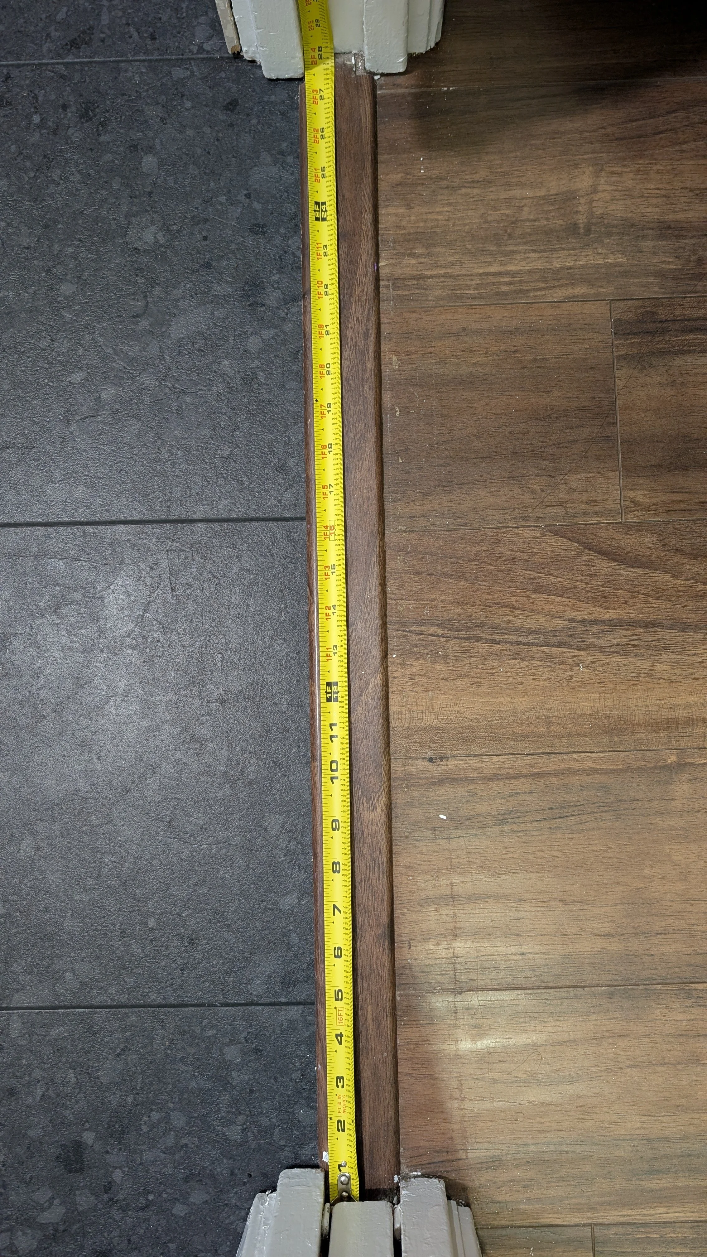 A measuring tape stretched across a wooden plank, with a dark tiled floor on one side and a wood floor on the other.