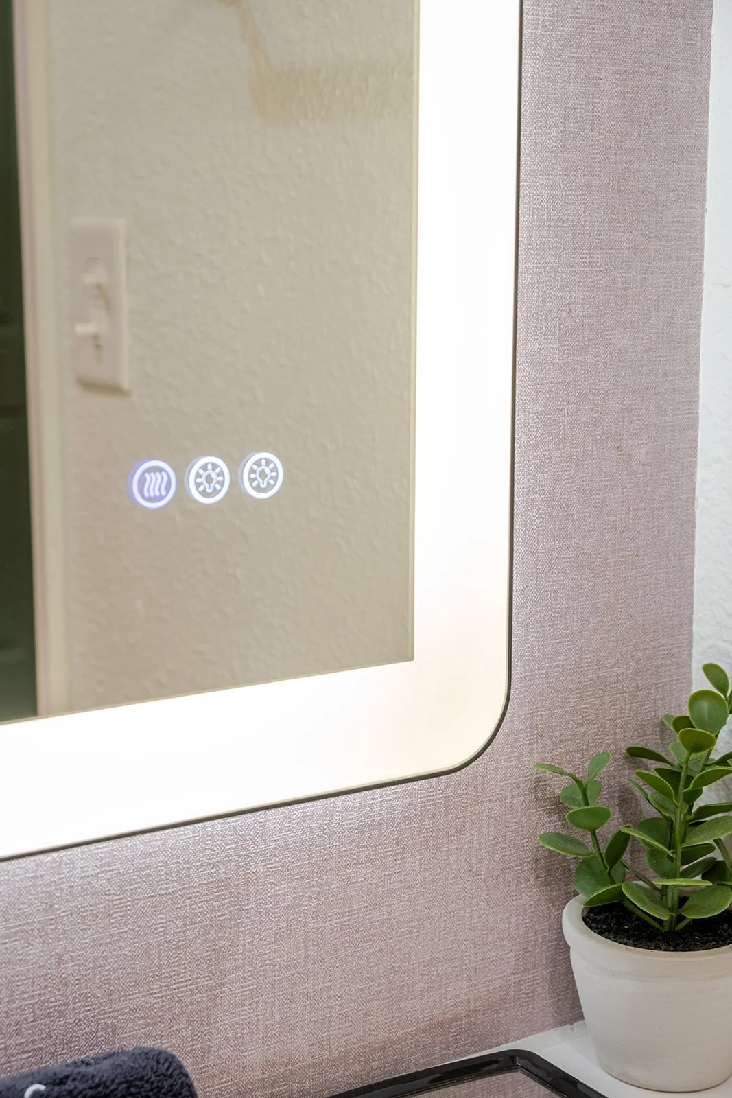 Close-up of a mirror with touch controls for heating and cooling, reflecting part of a wall and a potted green plant nearby.