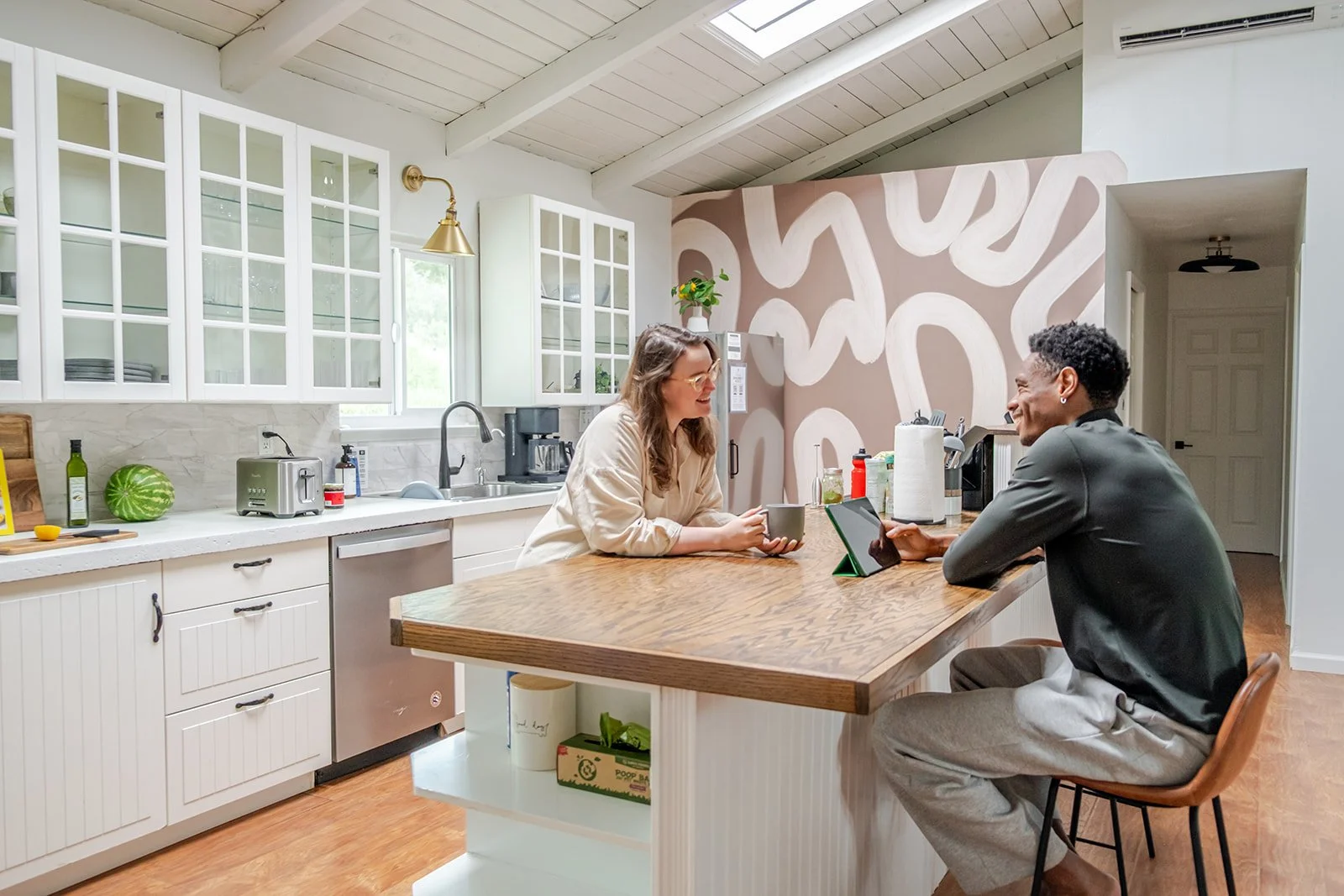 Two people sitting at a kitchen island, smiling at each other. The woman holds a mug, and the man uses a tablet. The kitchen features white cabinetry, a window, and a decorative wall with wavy patterns.