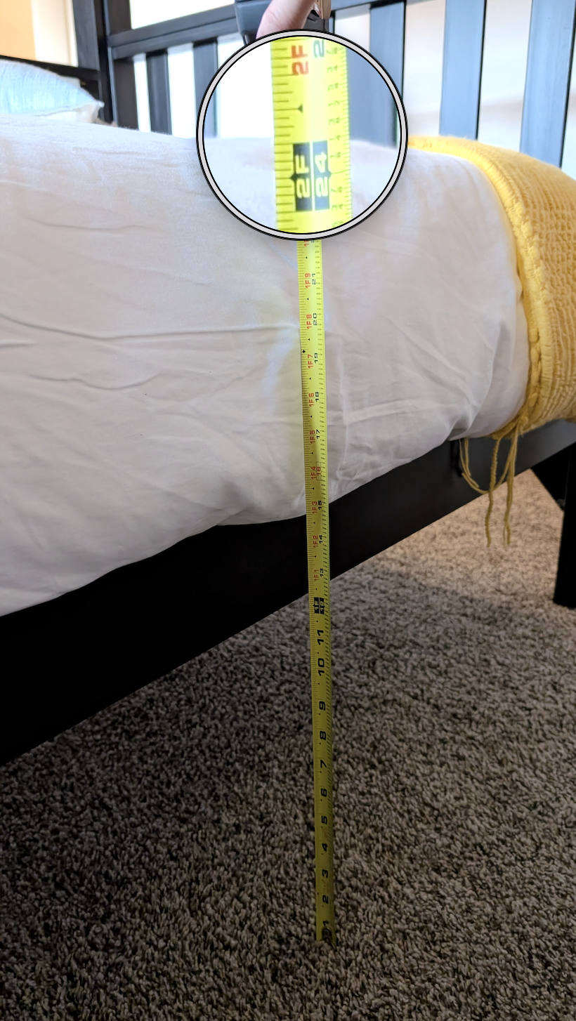 A yellow tape measure extended next to the side of a bed, showing measurement in inches. The tape displays about 24 inches.