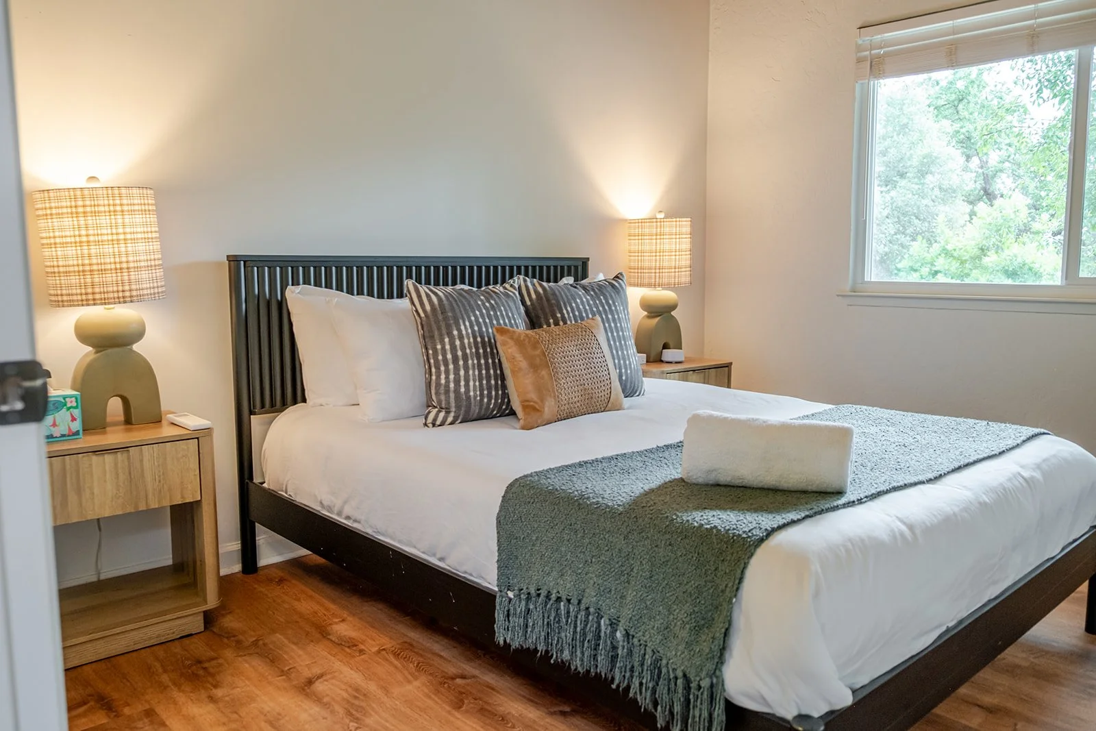 A neatly made bed with decorative pillows, a green throw blanket, and a rolled white towel, in a bedroom with wooden nightstands and lamps, near a window with view of green trees.