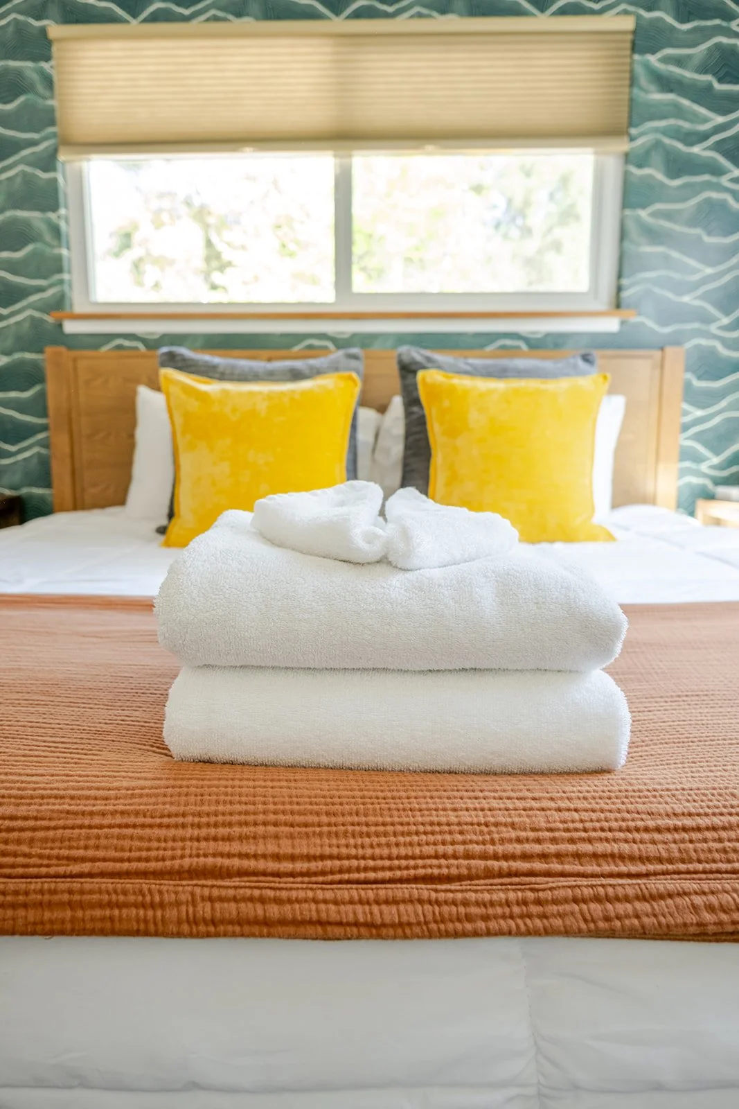 Stacked white towels with a folded towel on top shaped like a swan, on a bed with orange bedding, in a bedroom with a wooden headboard, yellow and gray pillows, and a window with beige blinds, decorative teal and white patterned wallpaper background.