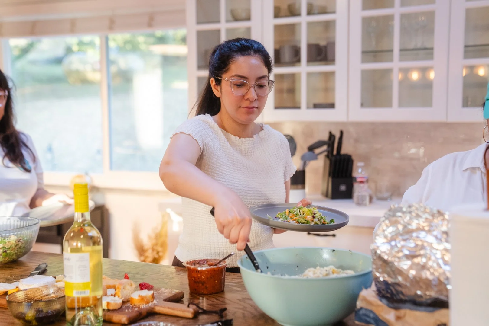 A woman preparing food in a kitchen, serving salad onto a plate, with various ingredients and utensils on the counter, including a bottle of wine, a bowl of salad, and a jar of salsa.