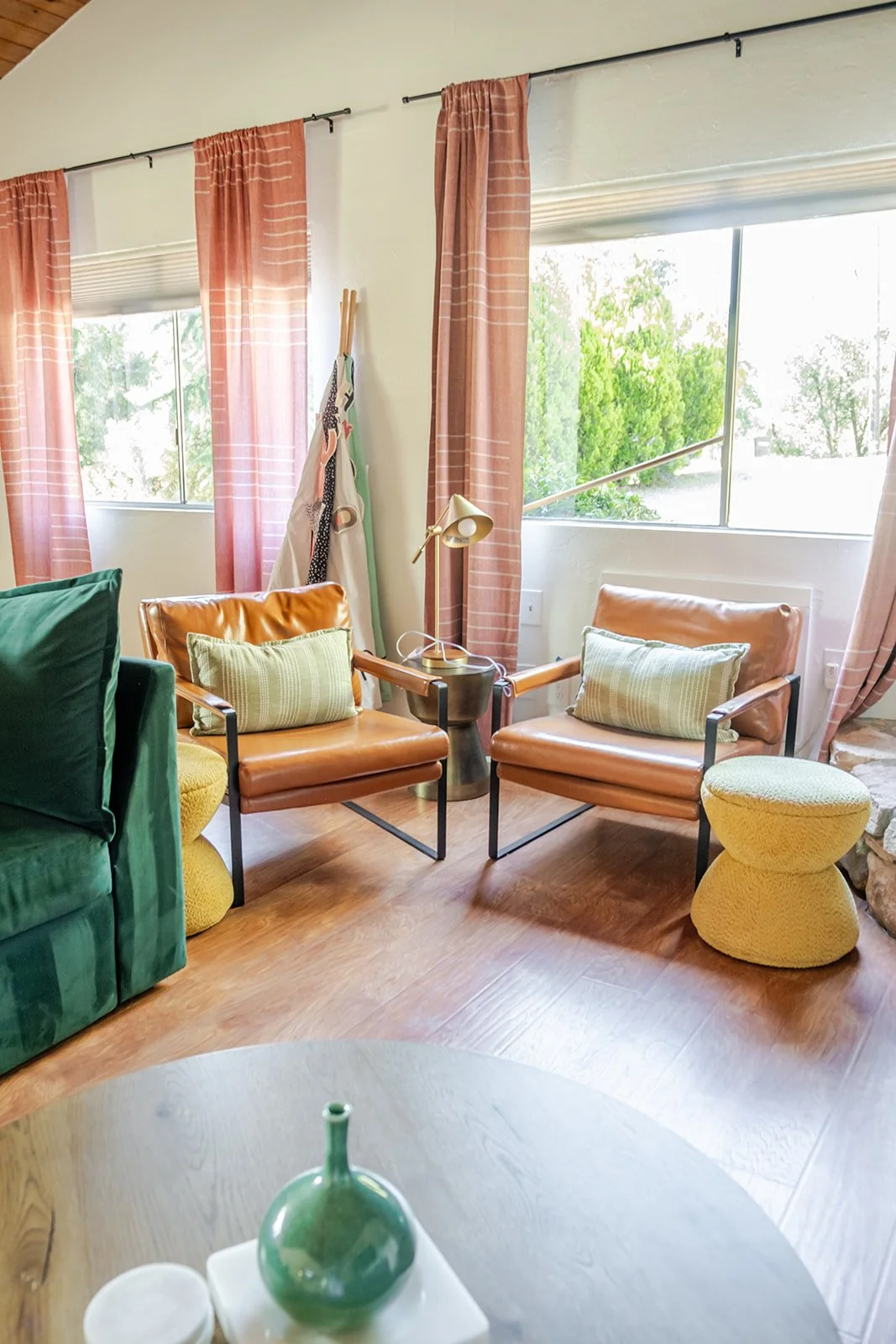 A cozy living room with two brown leather armchairs, a small black side table with a gold desk lamp, green and yellow cushions, pink curtains, large windows with a view of greenery, and a green velvet sofa partially visible on the left.