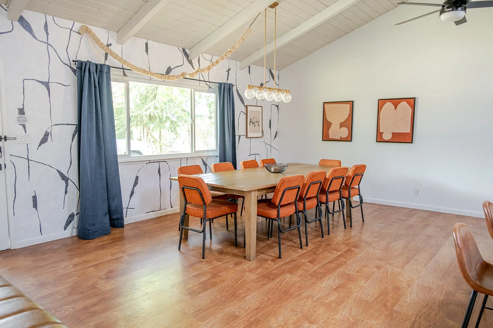 A dining room with a wooden table, eight orange upholstered chairs, a window with blue curtains, and artwork on the walls. The ceiling is vaulted with exposed white beams and a modern hanging light fixture. The floor is wood, and there is a ceiling f