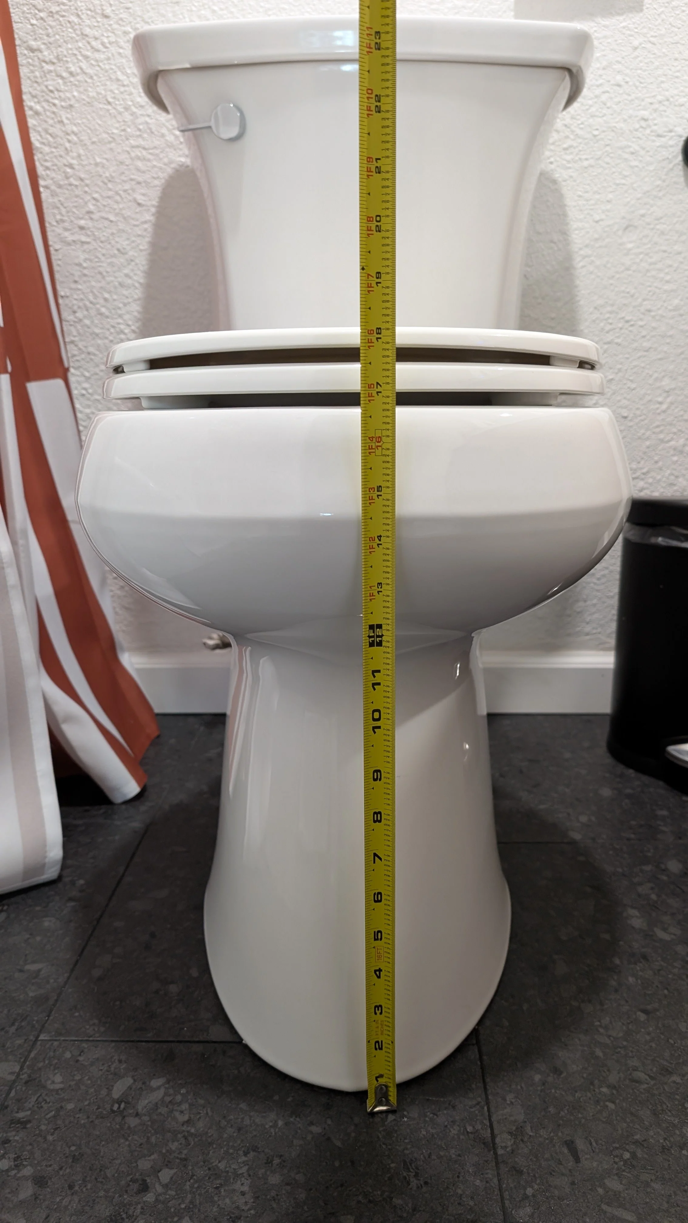 A white toilet with a yellow tape measure extended vertically from the floor to the top of the tank, measuring approximately 28 inches.