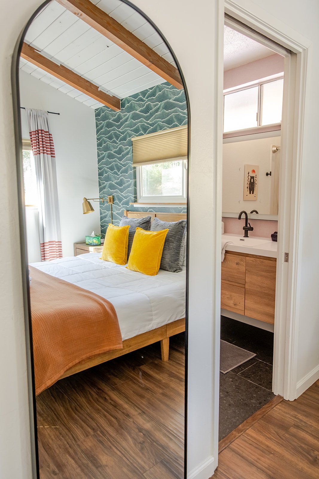 Reflection of a bedroom in a mirror shows a bed with gray pillows, two yellow pillows, and an orange blanket, with a window, a wooden bedside table, and a wall with patterned wallpaper. An attached bathroom with a vanity, sink, and art on the wall is