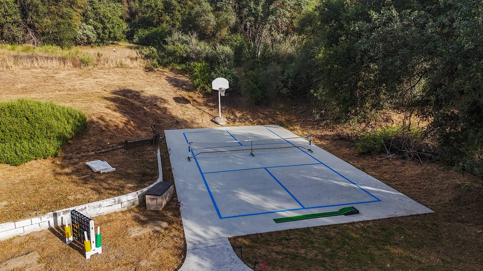 An outdoor sports court with blue lines and a net, a basketball hoop, and sports equipment, surrounded by trees and a small garden area.