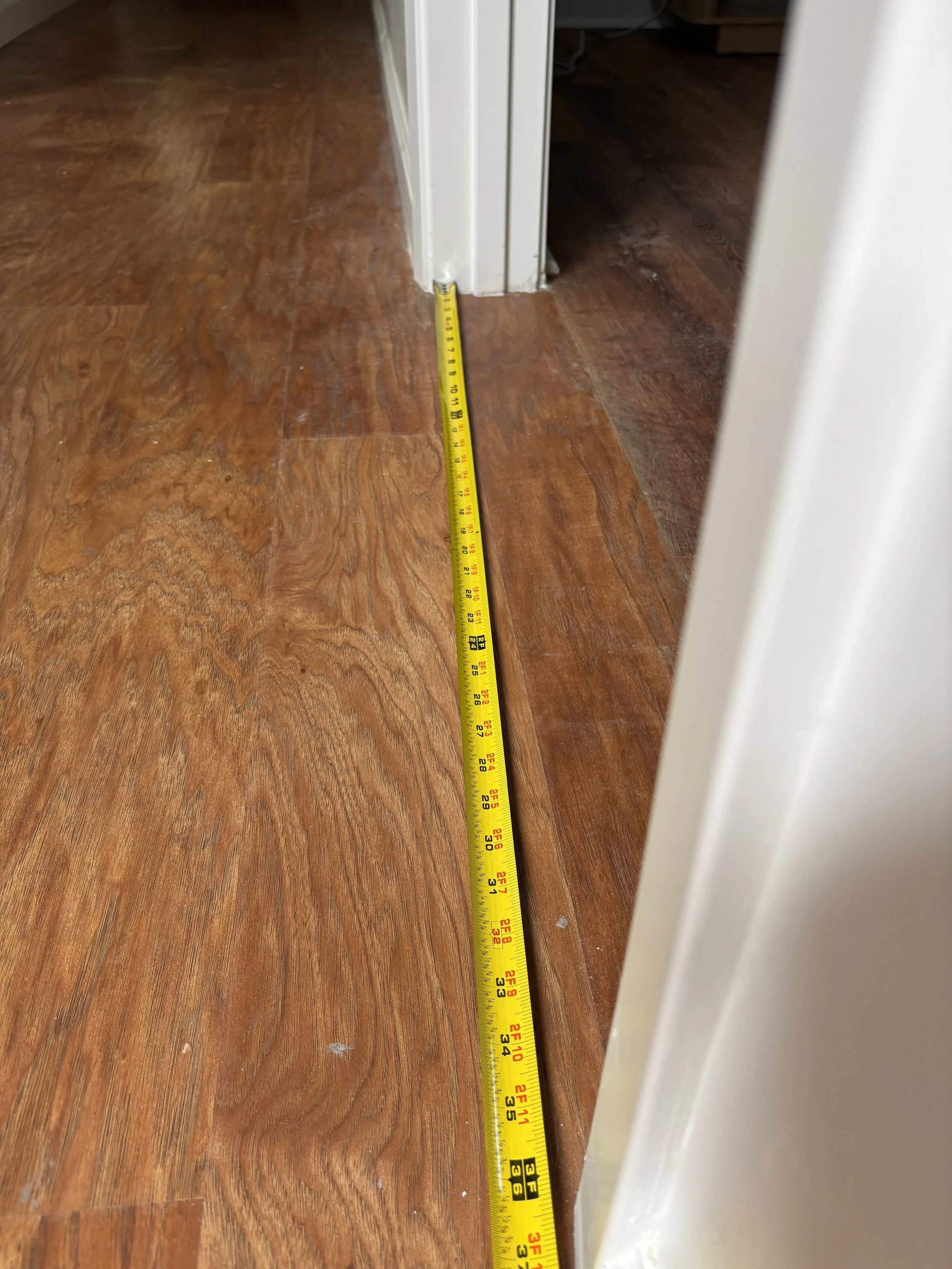 A yellow measuring tape extended along a hardwood floor, measuring approximately 36 inches, with a white door frame on the right side.