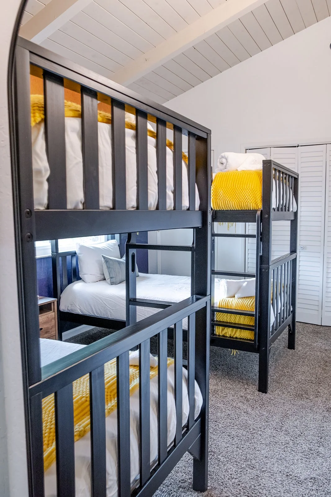 Room with bunk beds, showing top bunks with yellow blankets and rolled towels, and bottom bunks with white bedding and pillows, in a well-lit room with a sloped ceiling and closet doors.