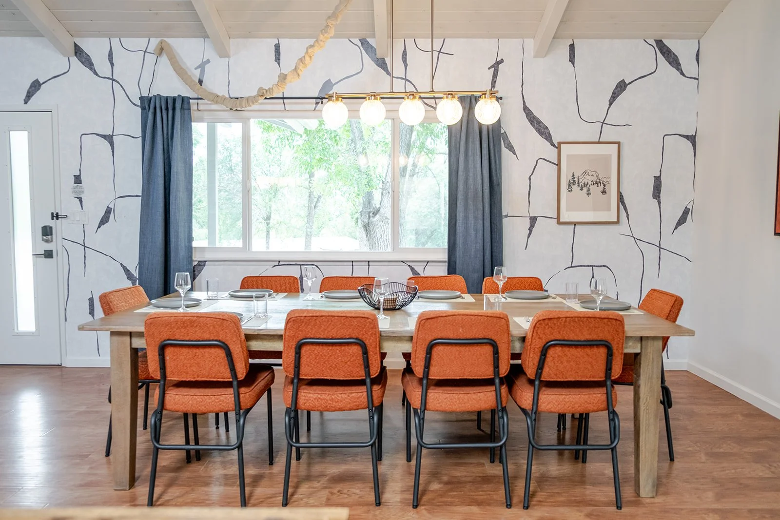 A dining room with a wooden table set with plates, glasses, and a wire basket, surrounded by orange upholstered chairs. A large window with dark blue curtains, a wall with black and white abstract design, and pendant lights hanging from the ceiling.
