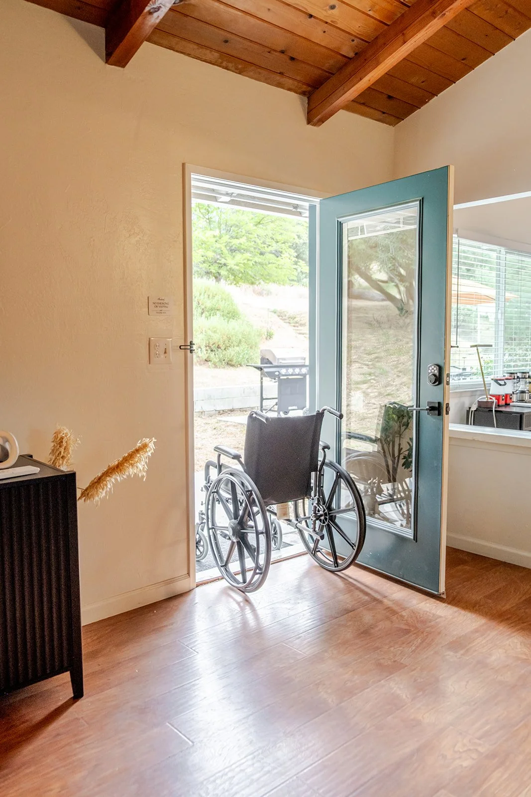 A wheelchair positioned inside a room, near a door leading outside to a garden with a barbecue grill. The room has wooden floors, beige walls, and a wooden ceiling.