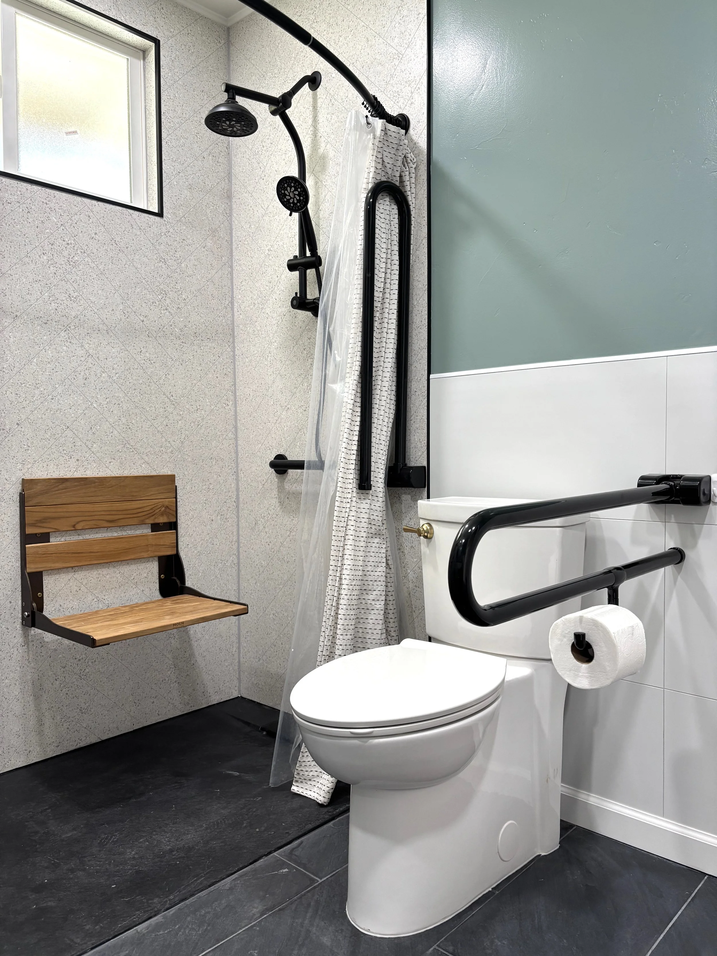 A disability-friendly bathroom featuring a white toilet with black support rails, a white and black grab bar, a toilet paper roll, a wooden bench, a shower with a black shower head, a curtain and a window, with beige textured walls and a black floor.