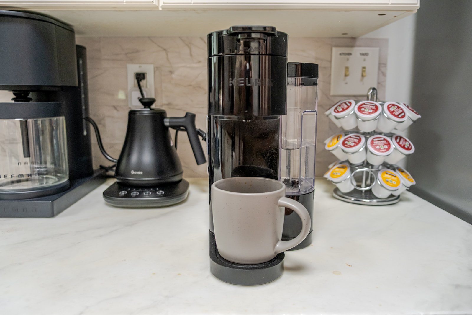 Coffee station with a coffee maker, a coffee grinder, a kettle, and a mug filled with coffee. K-Cup pods are arranged on a rotating stand in the background.
