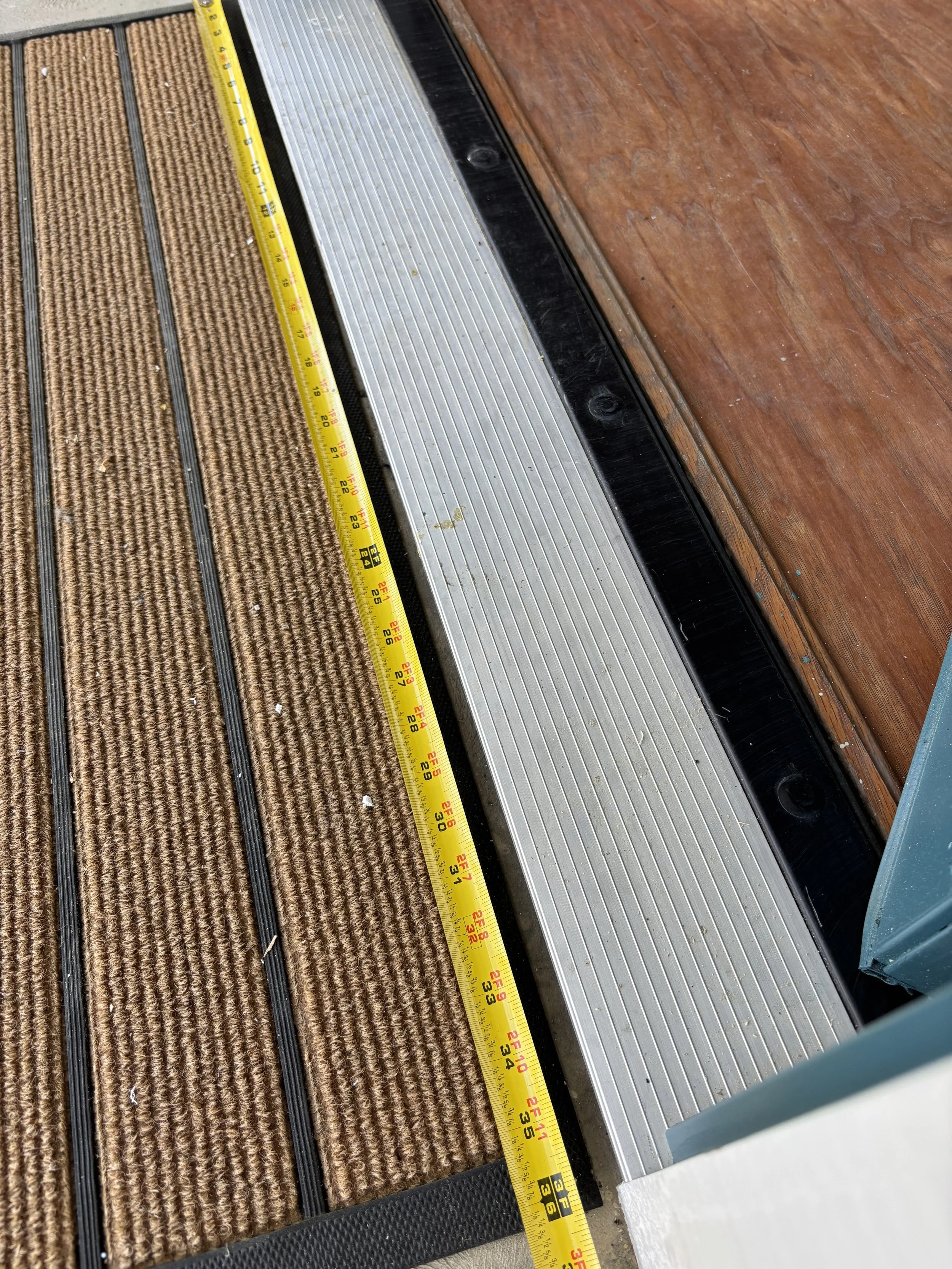 Close-up of a section of doormat and a metal threshold with a yellow measuring tape laid across them.