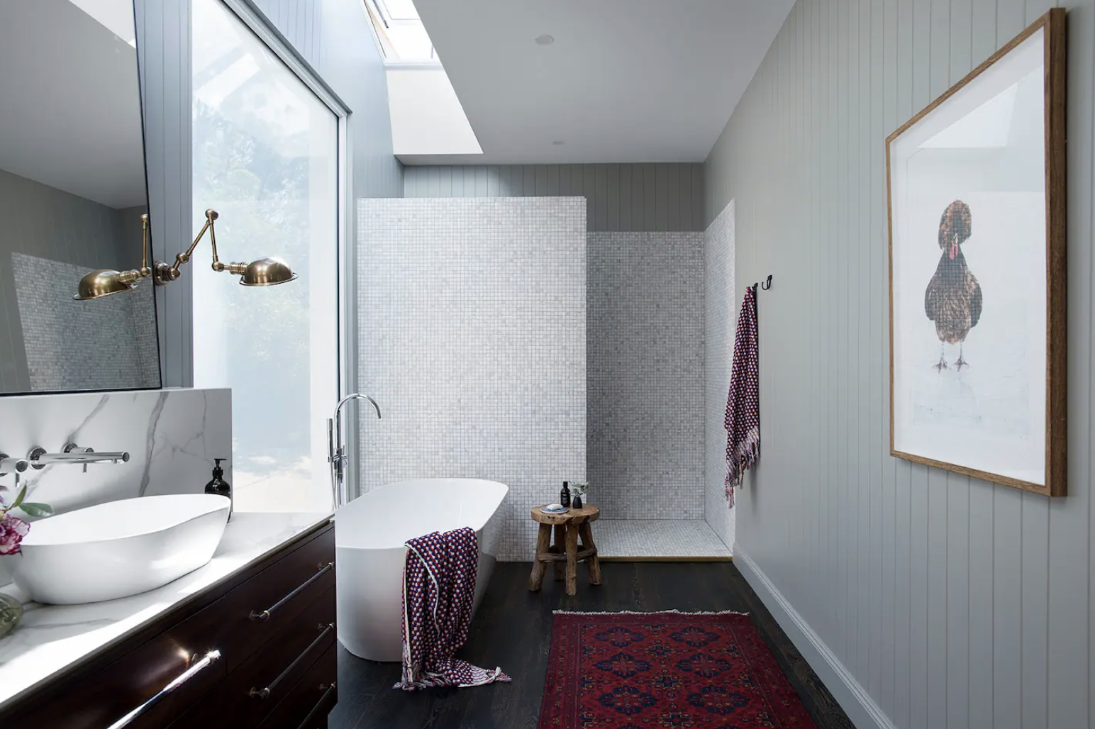 A modern bathroom with a white freestanding bathtub, a vessel sink on a dark wood vanity, a framed artwork of a turkey, and a walk-in shower with white mosaic tiles. Features include a skylight, large window, dark hardwood floor, and patterned red ru