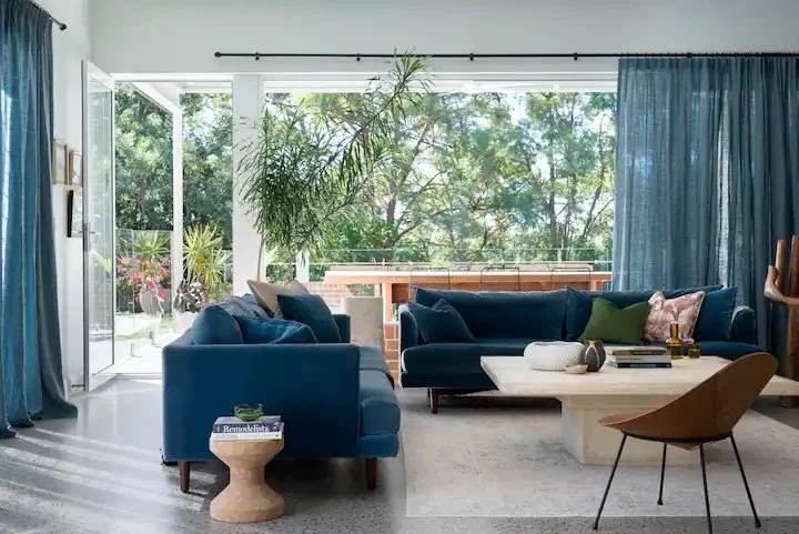 Living room with large window, blue sofas, a white coffee table, a brown accent chair, a tall plant, and a view of trees outside.