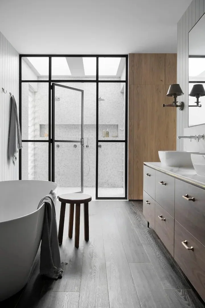 Modern bathroom with a freestanding bathtub, wooden stool, double sink vanity, glass enclosed shower area, large window, and dark wooden floor.