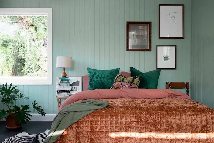 A cozy bedroom with a bed covered in a rust-colored velvet quilt, topped with green pillows and a floral pillow. On the left side, a small nightstand holds a white lamp and a stack of books. A large window on the left provides natural light, with a p