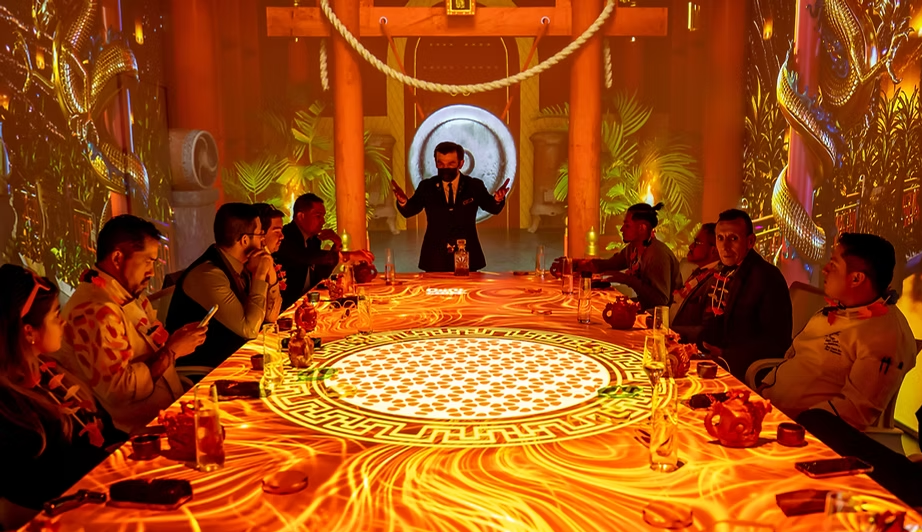 Un grupo de personas sentadas alrededor de una mesa larga en un ambiente decorado con temas orientales y dragones, con un presentador de pie en el centro de la mesa, en un escenario con fondos de fantasía y luces cálidas.