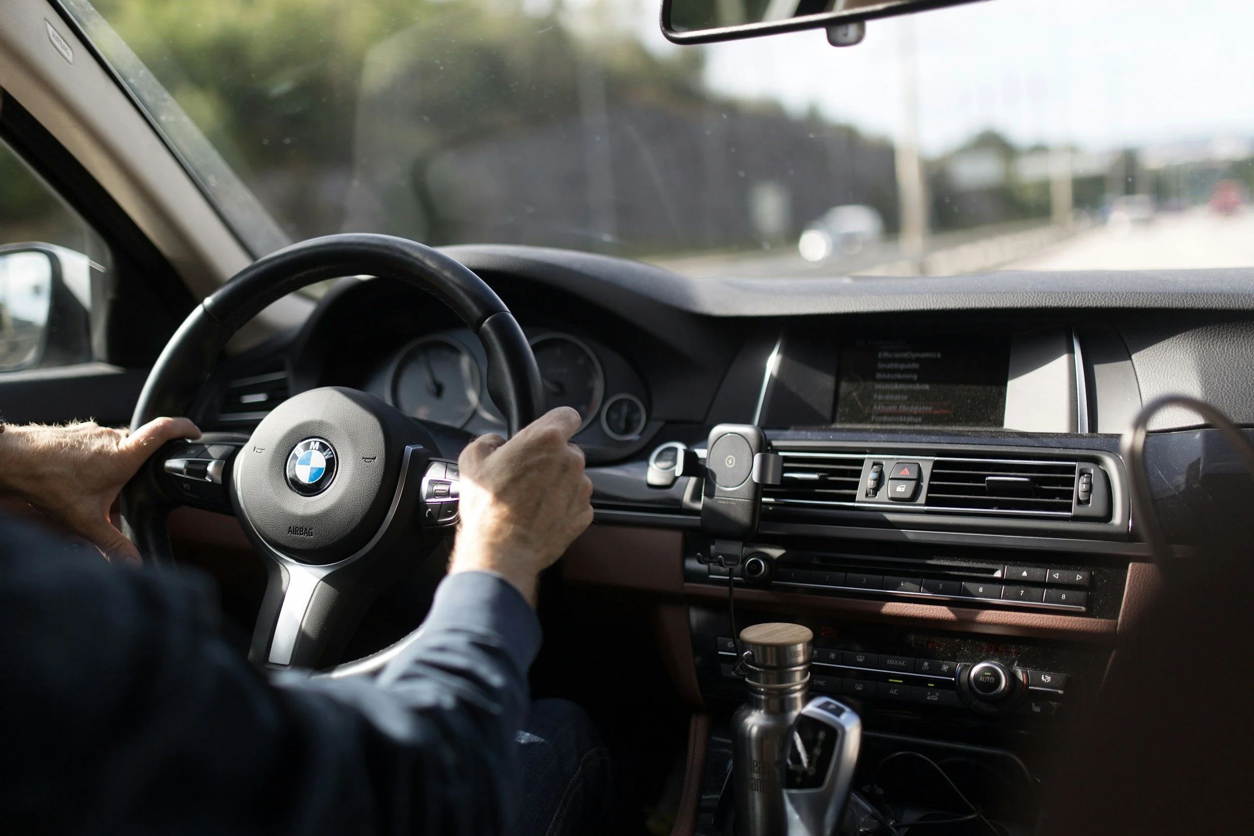 Vista del interior de un automóvil BMW desde el asiento del conductor, con las manos en el volante y una pantalla digital en el tablero, mientras conduce por una carretera con árboles y otros vehículos al fondo.