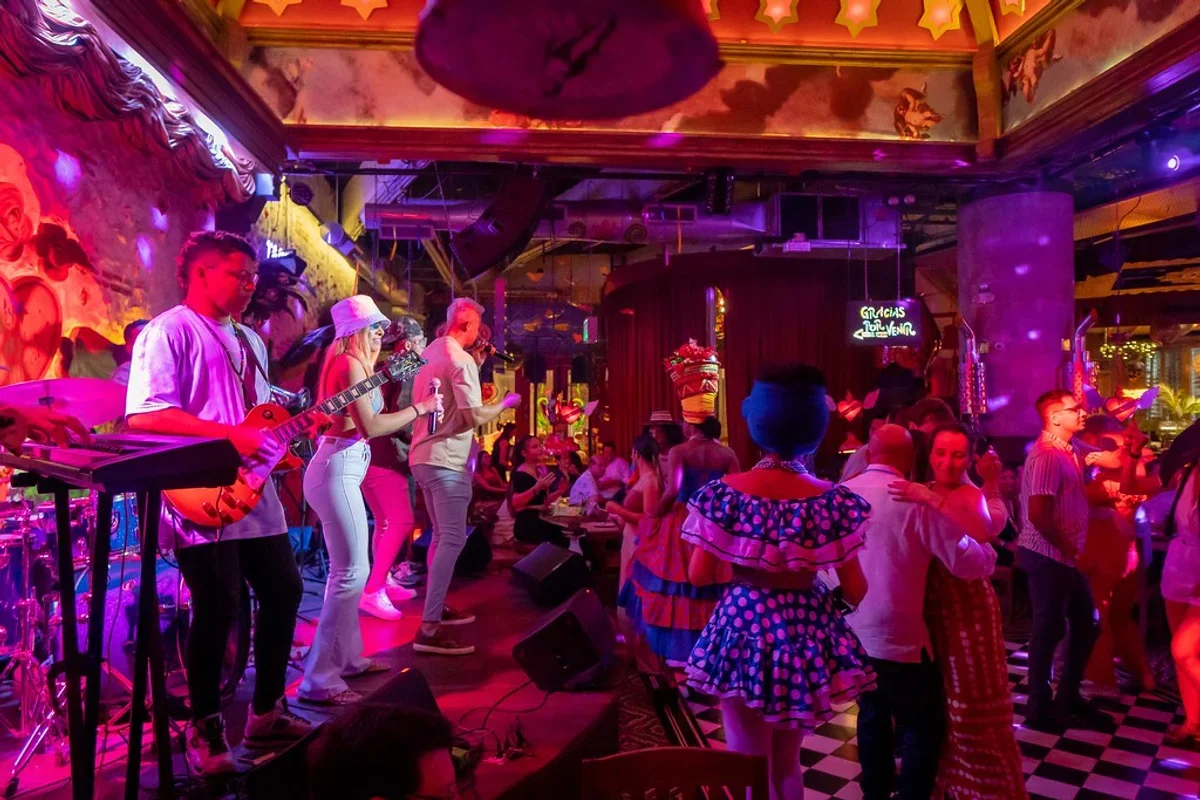 Gente bailando en un lugar con un escenario donde una banda toca música en vivo, luces de colores y decoraciones vibrantes.