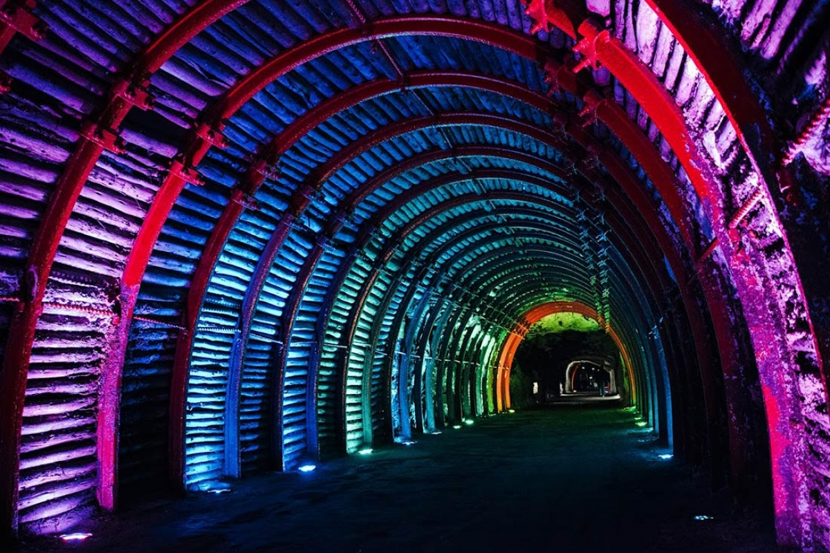 Túnel decorado con luces de colores en arcoiris, con paredes de hierro y madera, iluminado en tonos morados, azules, verdes y naranjas.