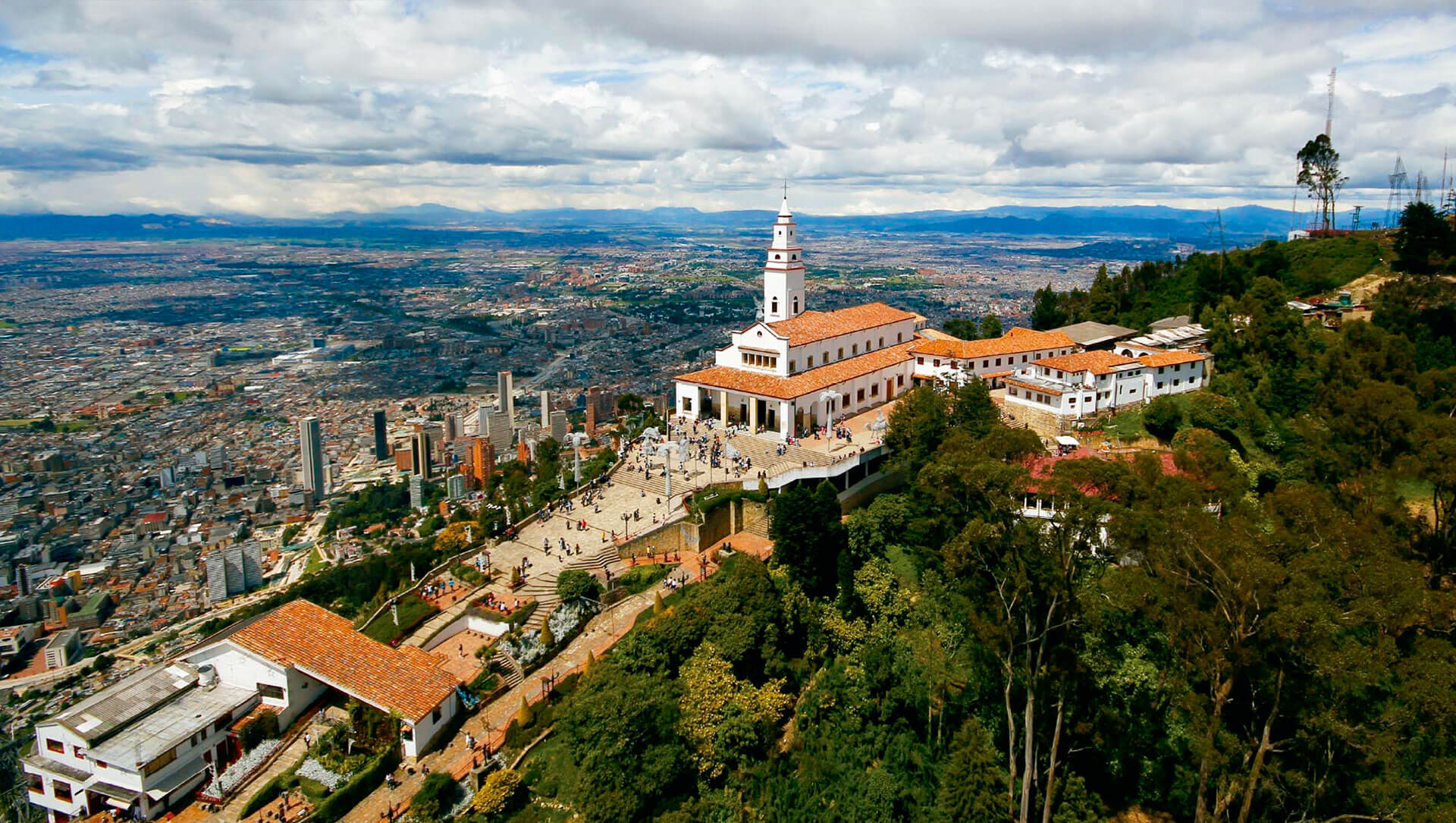 Vista aérea del Cerro del Cristo en la ciudad de Bogotá con la iglesia en la cima, rodeada de árboles y la ciudad en el fondo con edificios y montañas.