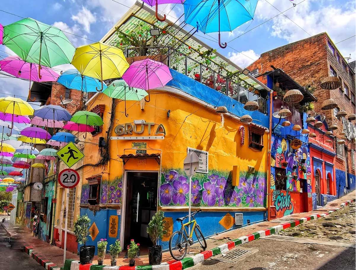 Una calle pintoresca con edificios coloridos, decoración de paraguas colgantes en el cielo, murales artísticos, un cartel de 'Gruta', una bicicleta y señalización vial.