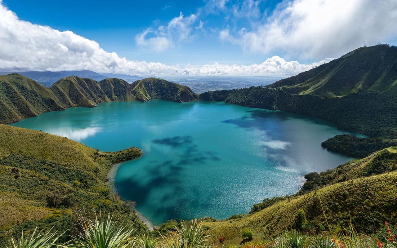 Lago rodeado de colinas verdes y montañas bajo un cielo con nubes.
