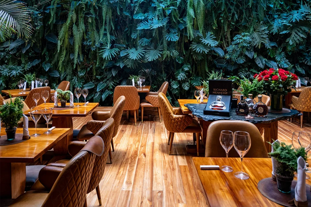 Sala de restaurante elegante con mesas de madera, sillas acolchadas y un fondo de pared cubierta de plantas verdes.