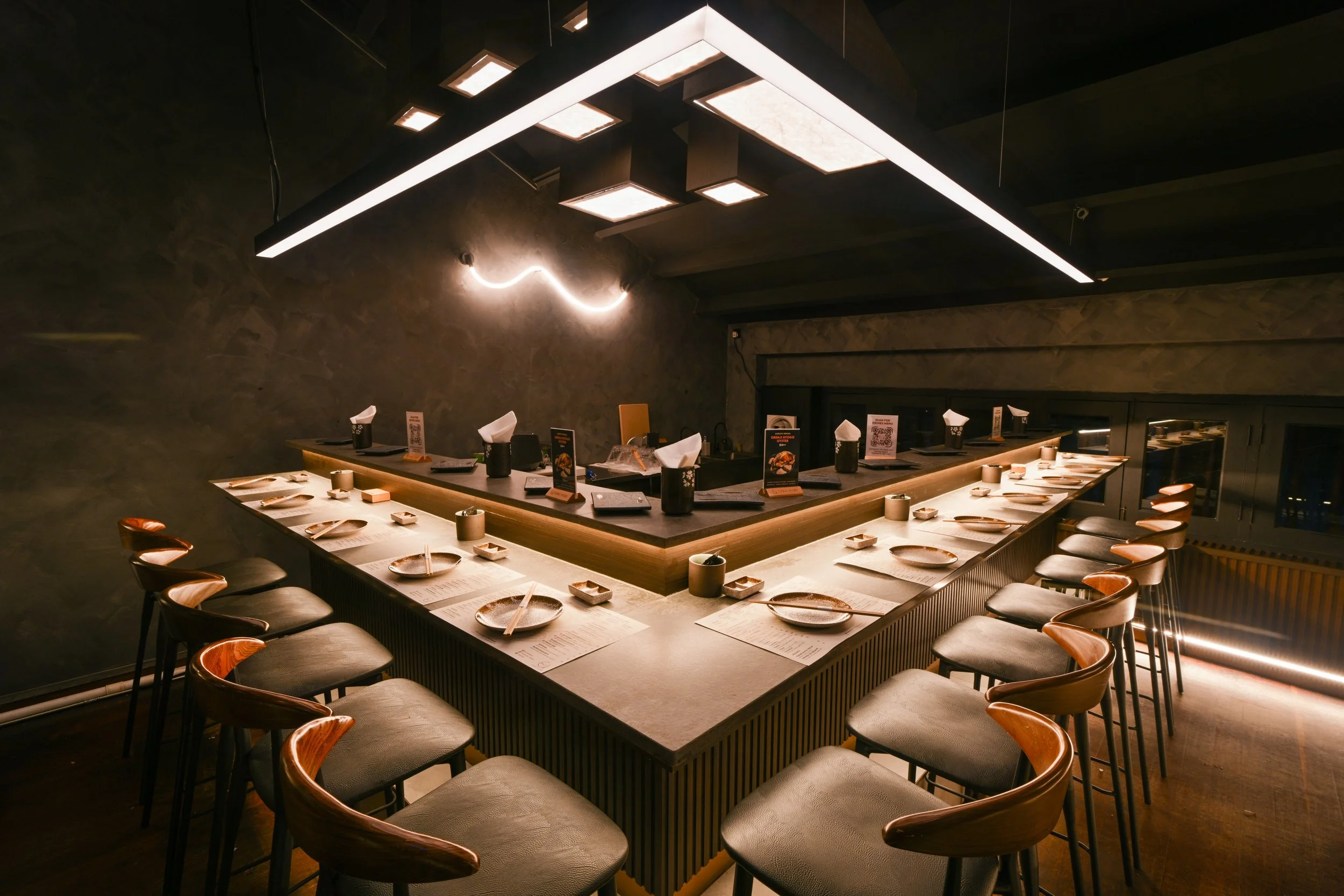 Empty modern Japanese restaurant with a U-shaped counter, seating for numerous guests, menus, chairs, and tableware, under contemporary lighting fixtures.