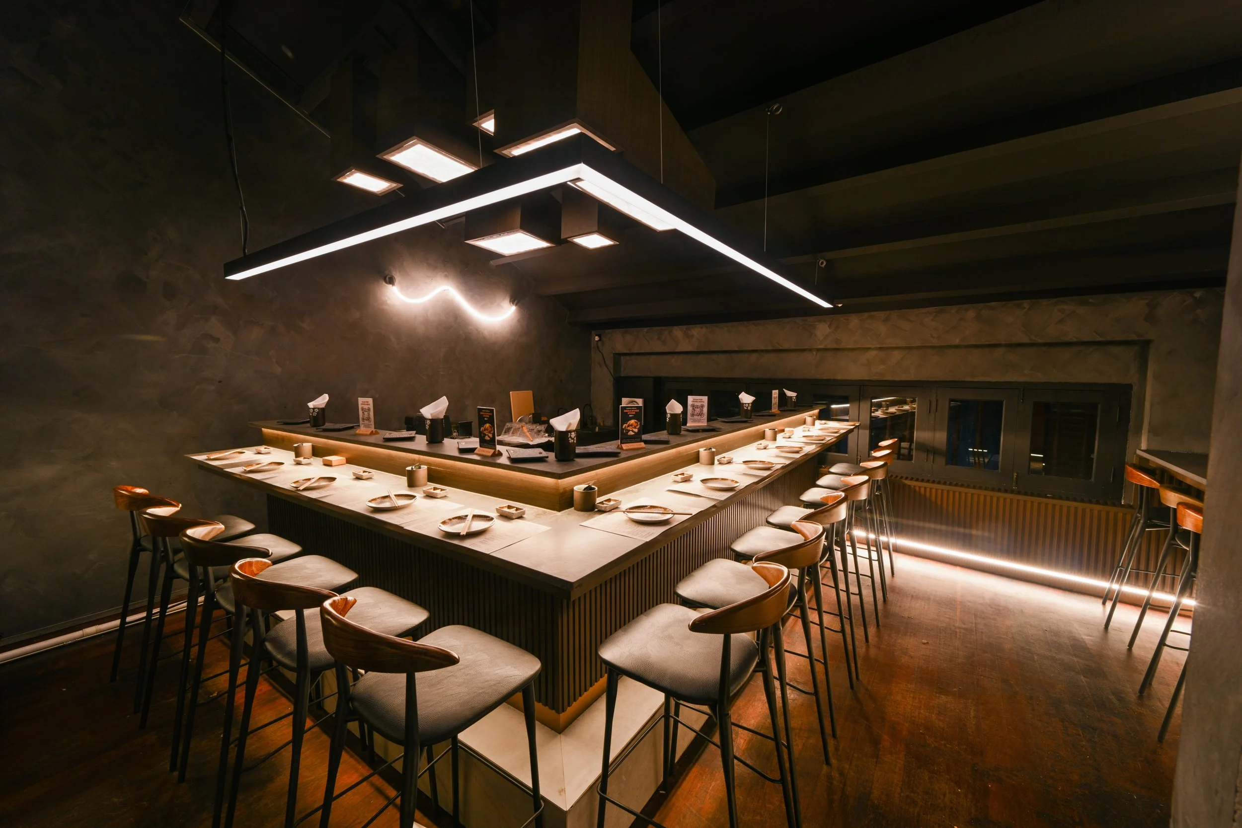 Empty modern sushi bar with a U-shaped counter, dinner plates, small cups, and chopsticks, illuminated by a sleek rectangular hanging light fixture in a dimly lit room.