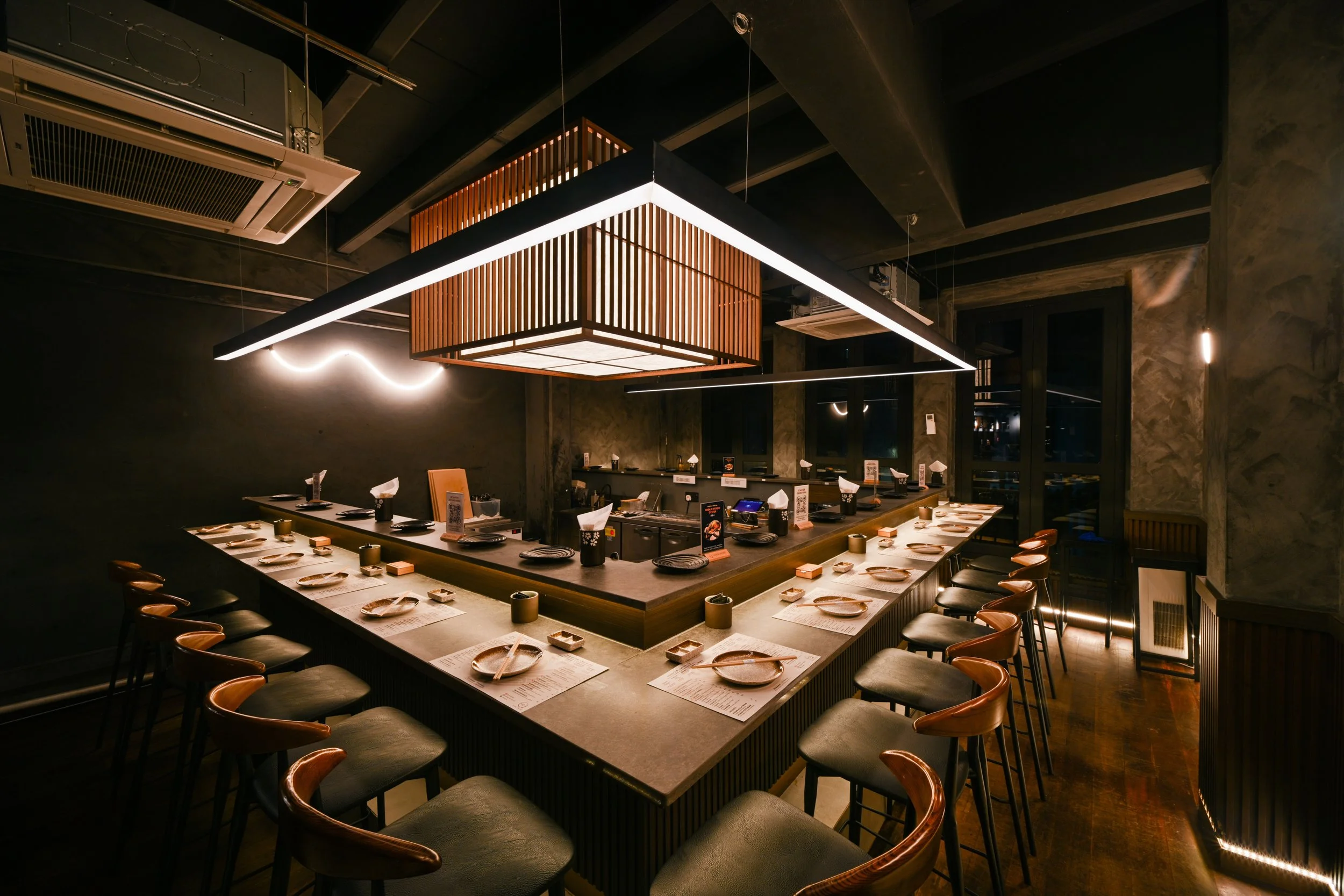 Interior of an upscale sushi restaurant with a U-shaped counter set with plates, napkins, and utensils, illuminated by modern lighting fixtures.