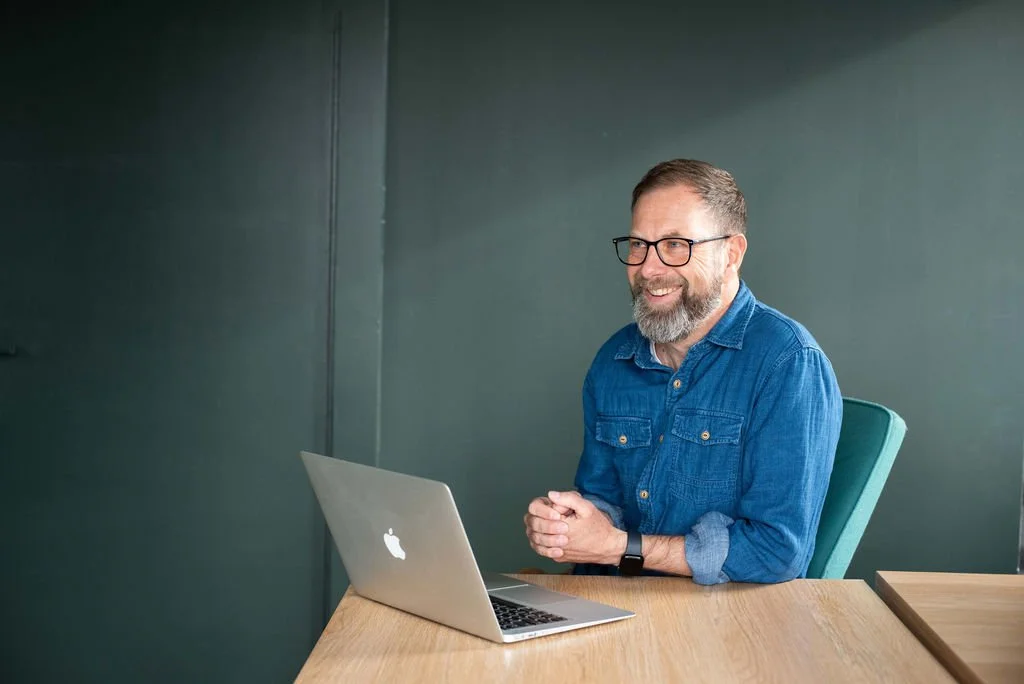 A smiling man with glasses and a beard sitting at a wooden desk with a silver laptop in front of him, in a room with dark green walls.